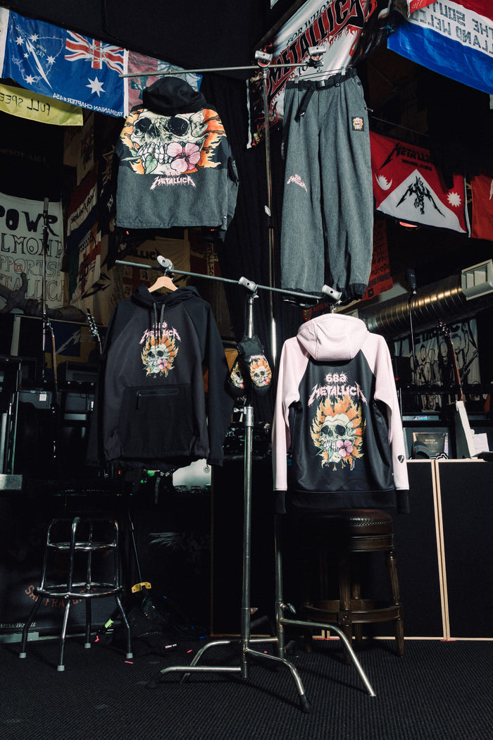 Two 686 Women’s Metallica Burning Skull Insulated Jackets and a pair of pants featuring a graphic of a flaming skull with flowers hang on a clothing rack in a music-themed room.