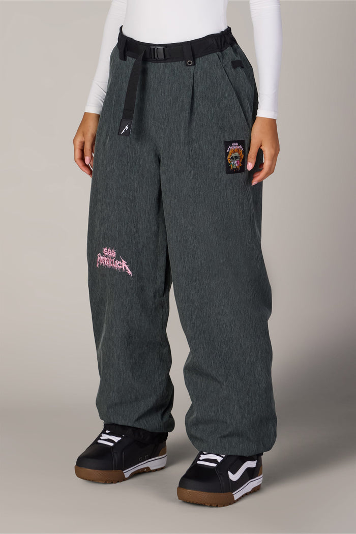 686 Women’s Metallica Burning Skull Shell Pant in dark grey, featuring a waterproof woven design, a burning skull graphic on the thigh, a small hip patch, and a black webbed belt at the waist.