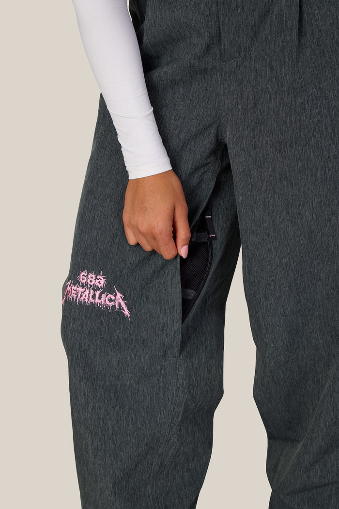 Close-up of 686 Women’s Metallica Burning Skull Shell Pant, featuring waterproof woven charcoal gray fabric, a pink Metallica logo embroidered on the right thigh, and an open side zipper pocket.