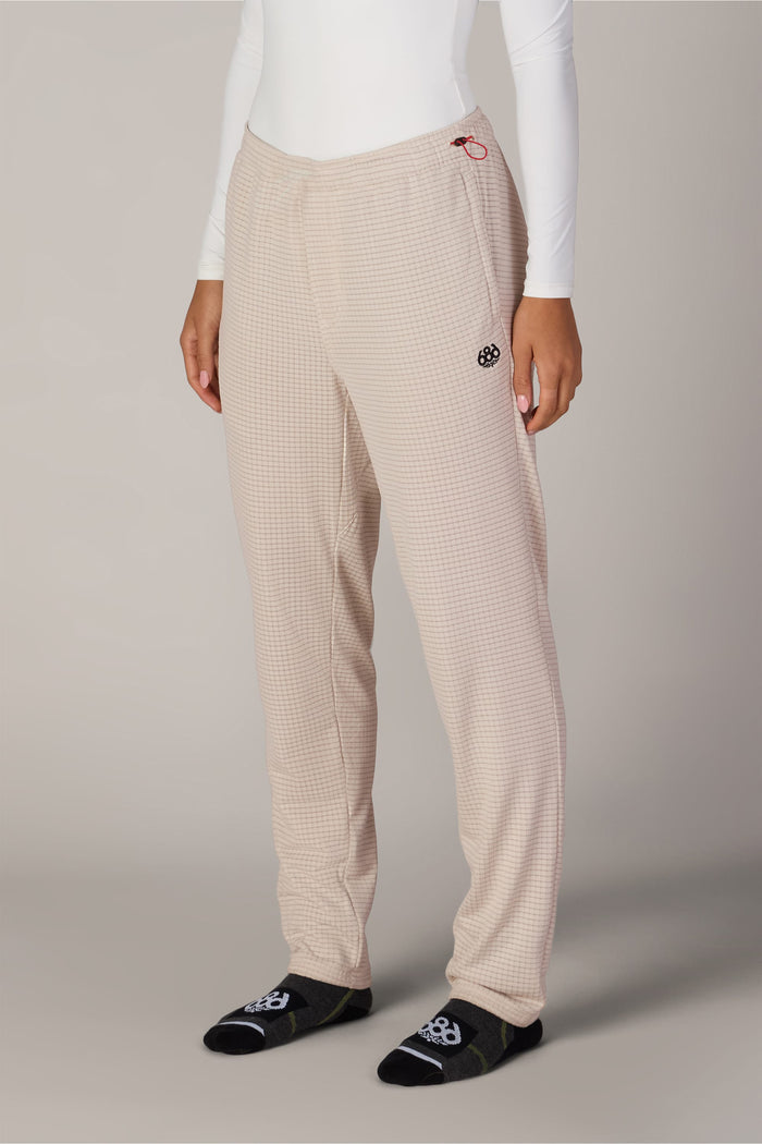 A person is shown from the waist down, wearing the 686 Women’s Mission Grid™ Pant by 686 in a beige checkered pattern, paired with a white long-sleeve top and black socks featuring white designs, standing against a plain background.