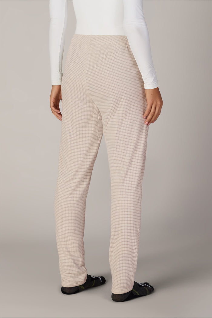 A person stands facing away, wearing 686 Women’s Mission Grid™ Pants by 686 in a beige checkered pattern, paired with a white long-sleeve shirt and black socks. The background is plain and light-colored.
