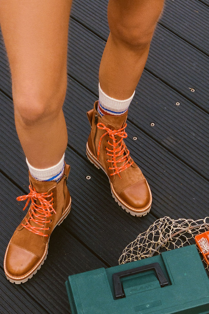 Person wearing brown boots with orange laces on a wooden floor, next to a green toolbox.