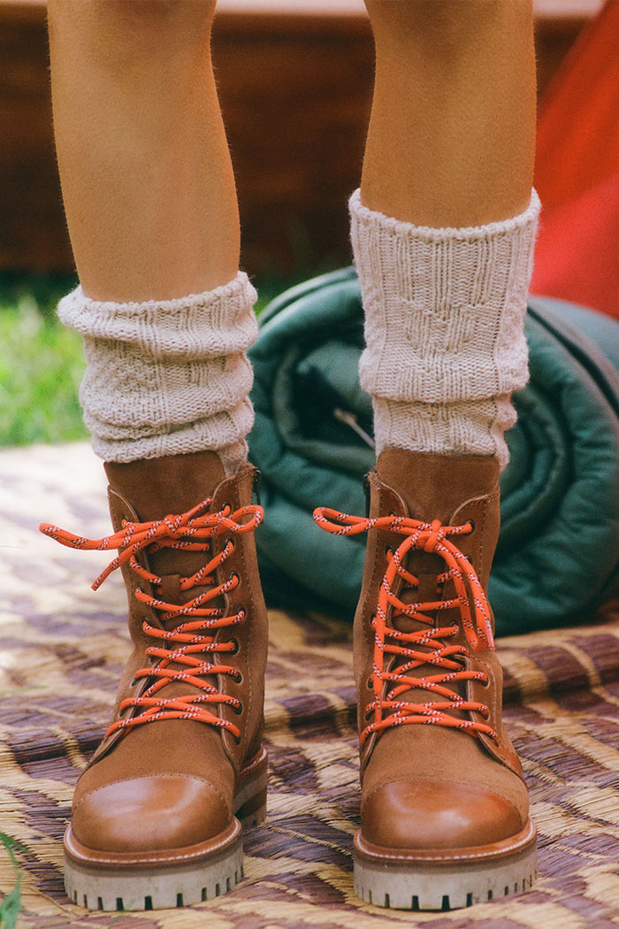Brown boots with orange laces worn with white socks on a patterned surface.