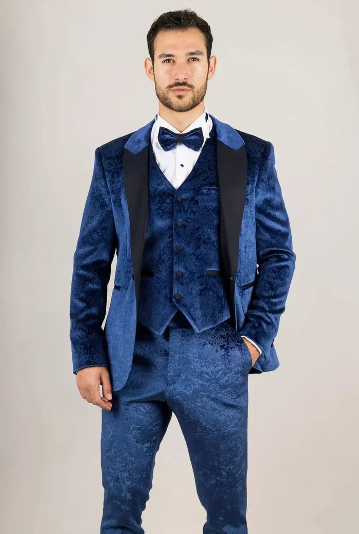 Man wearing a blue velvet tuxedo suit with black lapels, matching vest, white shirt, and bow tie, standing against a plain background.