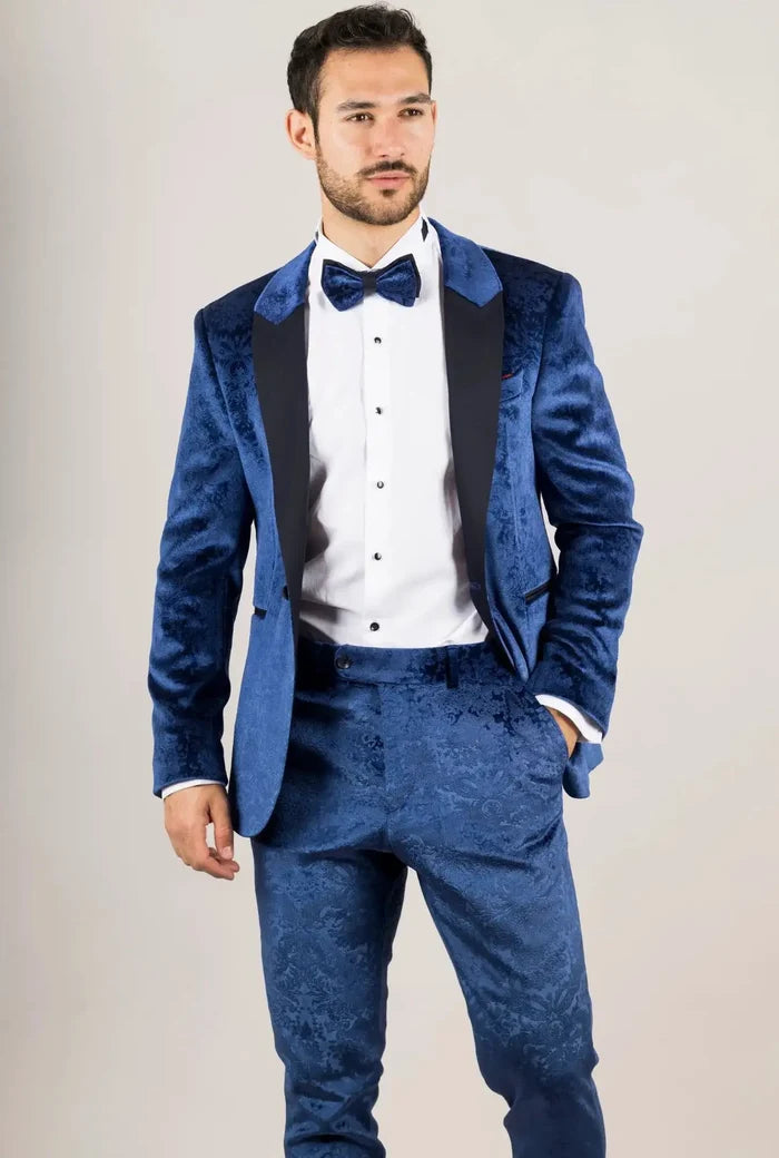 Close-up of a man in a blue velvet tuxedo with a detailed floral pattern, white shirt, and bow tie, looking serious.