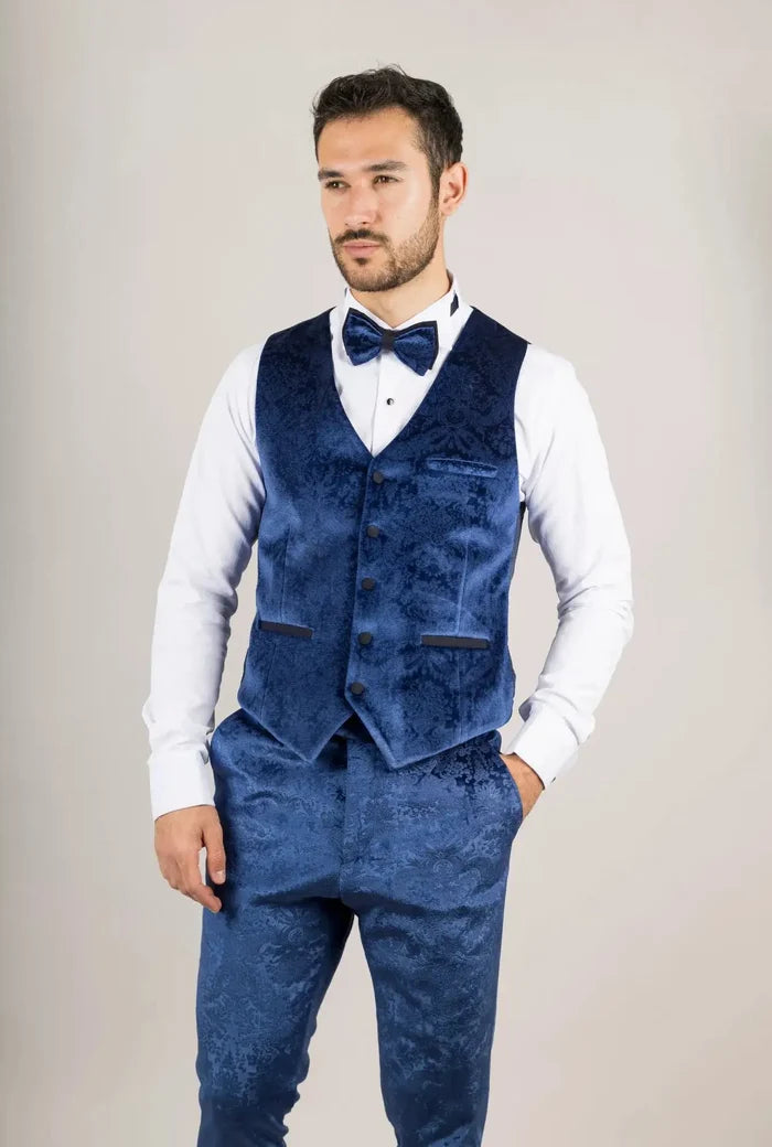 Man in a blue velvet tuxedo with a floral pattern, white shirt, and bow tie, standing with hands in pockets, facing forward.