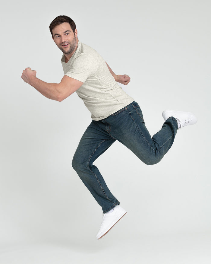Man wearing cream-colored short-sleeve t-shirt and dark blue athletic-fit jeans with white sneakers, jumping in dynamic pose.
