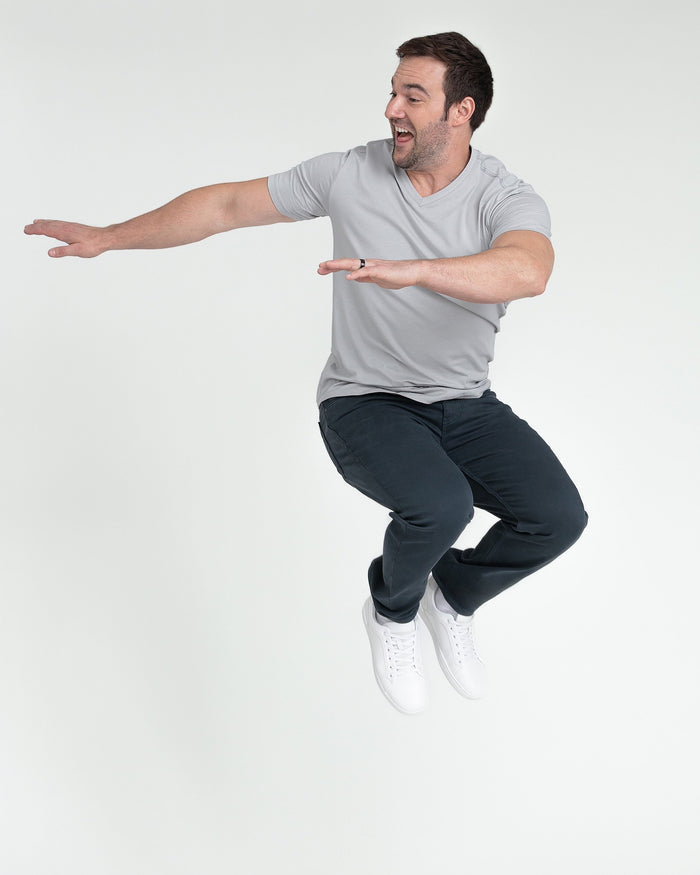 Man wearing gray v-neck t-shirt and dark slate athletic fit pants with white sneakers, jumping with arms extended.