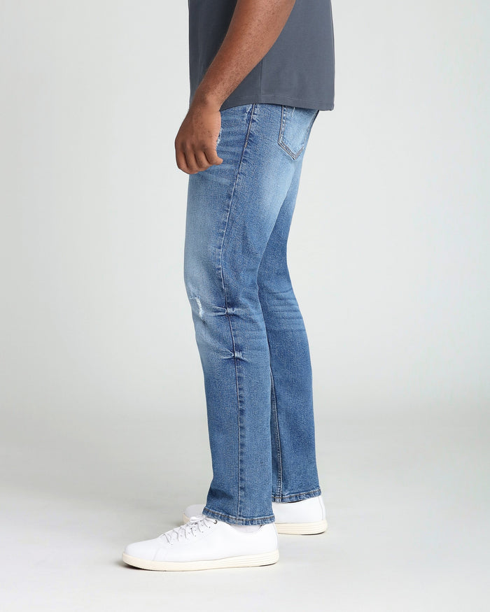 Men's medium blue athletic fit jeans with ripped details at the thigh, worn with white sneakers.