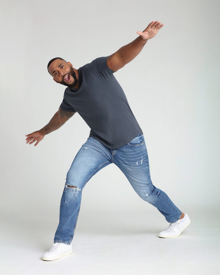 Man wearing dark gray t-shirt and medium blue ripped jeans with white sneakers in dynamic pose.