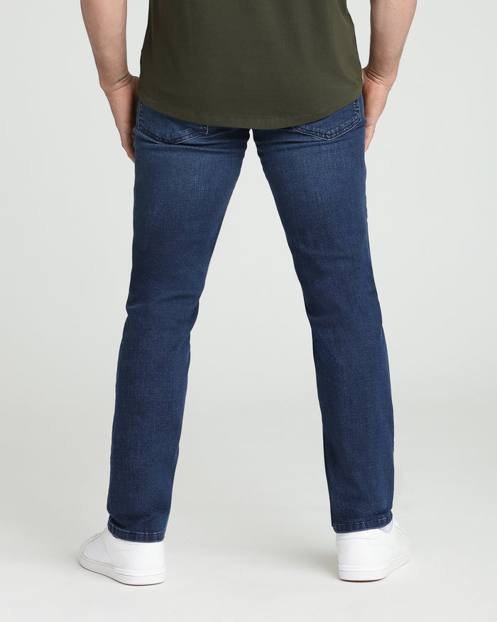 Man wearing olive green t-shirt and dark blue athletic fit jeans with white sneakers, shown from behind.