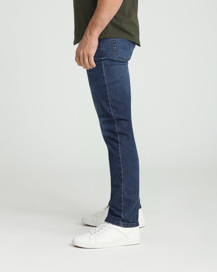 Man wearing dark blue athletic fit jeans with olive green shirt and white sneakers, shown from the side.