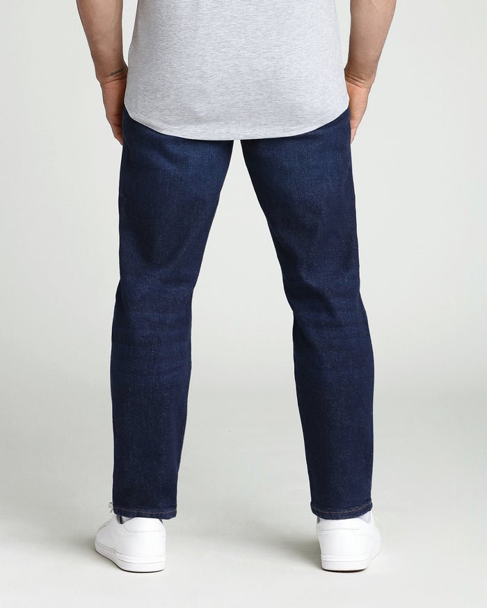 Man wearing light gray t-shirt and dark blue athletic fit jeans with white sneakers, shown from behind.