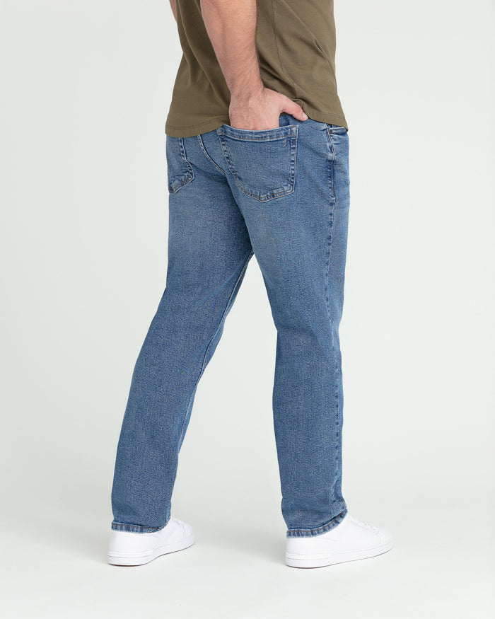 Medium blue athletic fit jeans on model wearing olive shirt and white sneakers, shown from side angle.