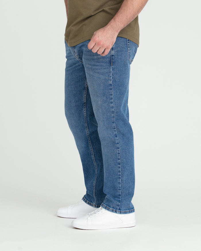Man wearing medium blue athletic fit jeans with olive shirt and white sneakers, shown from waist down in side profile.