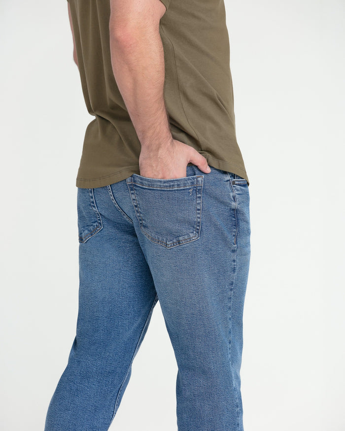 Man wearing medium blue athletic fit jeans with a khaki shirt, showing side view of waistband and pocket detail.