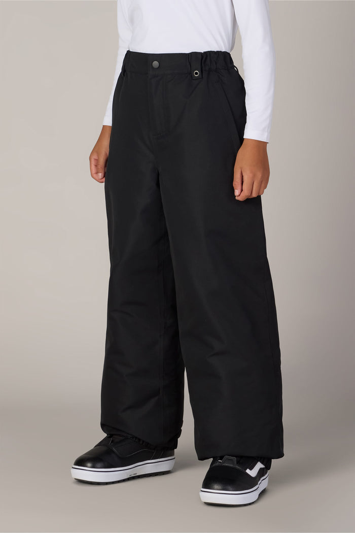 A child wearing the 686 Boys’ Dojo® Insulated Pant in black, a white long-sleeve shirt, and black-and-white sneakers stands against a plain light gray background.