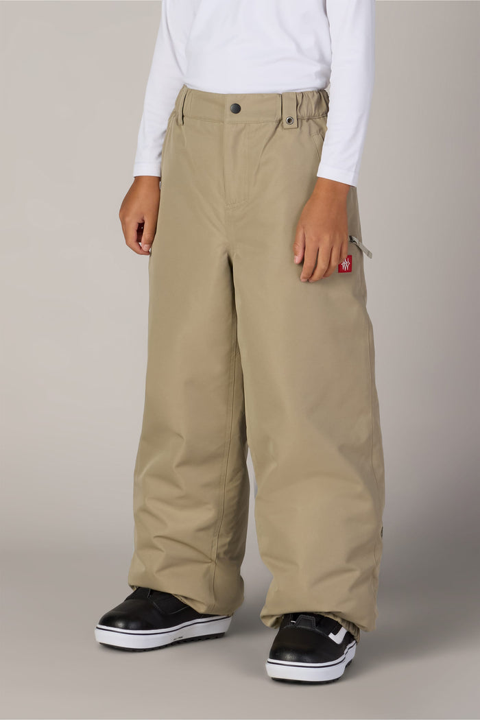 A child wearing a white long-sleeve shirt, the 686 Boys’ Dojo® Insulated Pant in khaki, and black sneakers with white soles stands against a plain light gray background.