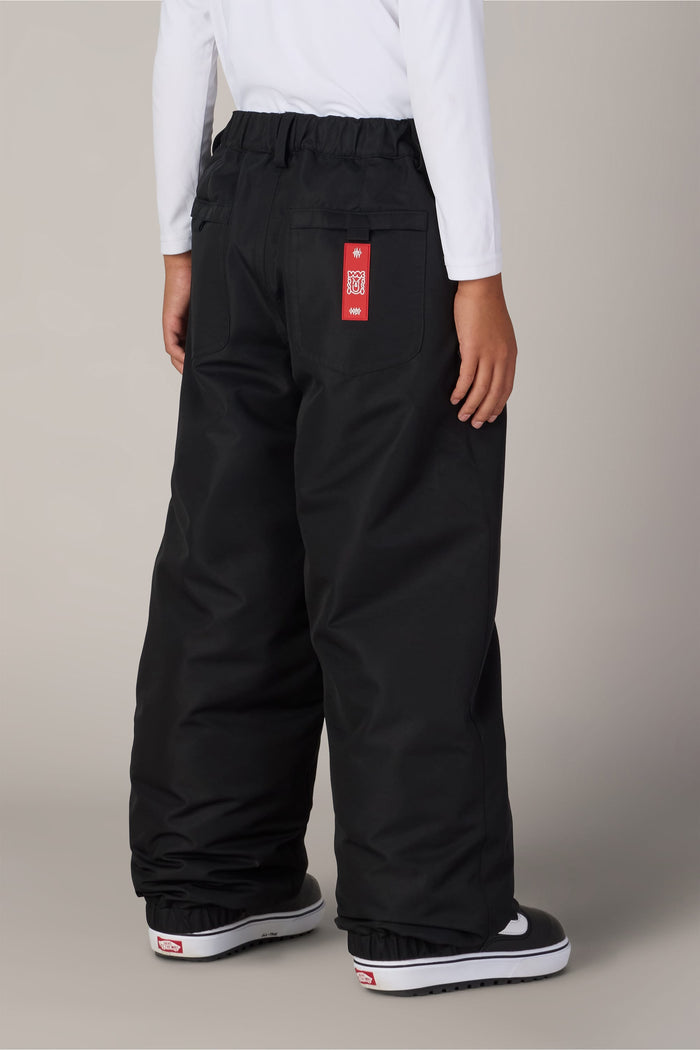 A child wearing the Boys’ 686 Dojo® Insulated Pant in black, featuring a red label on the back pocket, pairs it with a white long-sleeve shirt and black-and-white shoes while standing facing away from the camera against a plain background.
