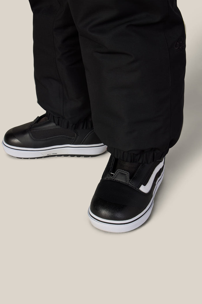 A close-up of a person wearing the 686 Boys’ Dojo® Insulated Pant and black slip-on sneakers with white soles and a white stripe design, standing on a light-colored floor.