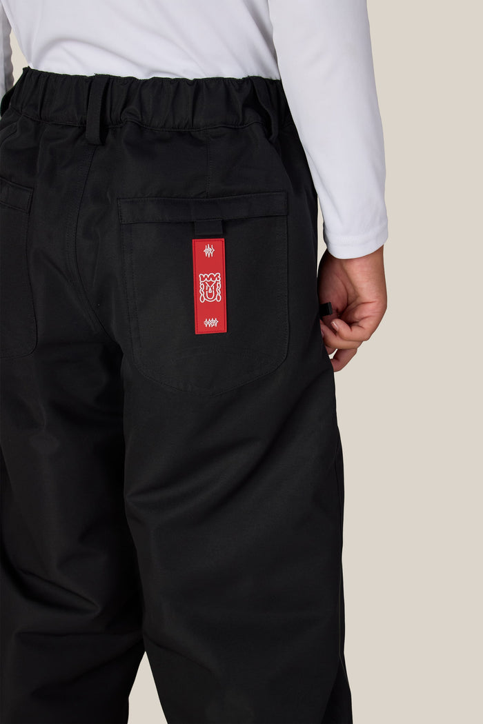 A boy is shown from the back wearing black 686 Dojo® Insulated Pants and a white long-sleeve shirt. The pants feature a red tag with white symbols on the back right pocket.
