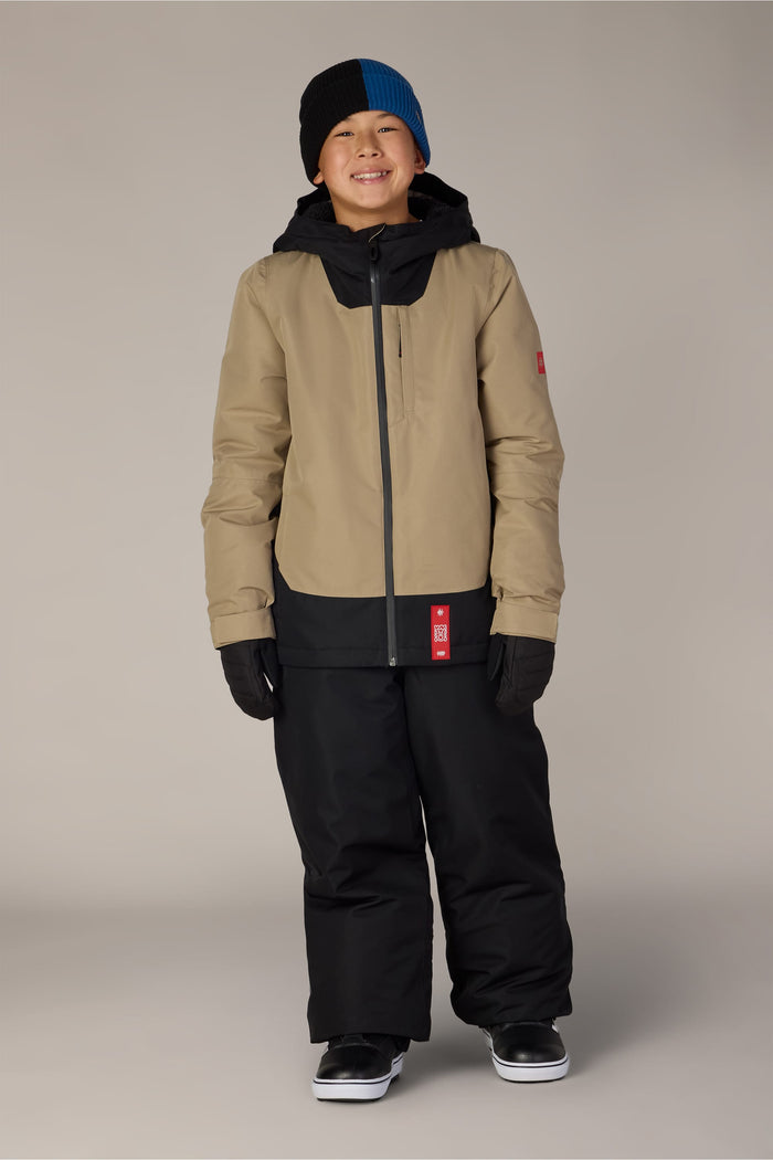 A smiling child wearing the 686 Boys’ Dojo® Insulated Pants in black, paired with a beige and black winter jacket, black gloves, a black and blue beanie, and black sneakers, standing against a plain background.