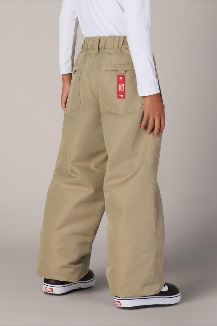 A person is wearing the Boys’ 686 Dojo® Insulated Pant by 686—loose, wide-legged khaki pants with a red tag on the back pocket—paired with a white long-sleeve shirt and black slip-on shoes with white soles, standing on a light background.