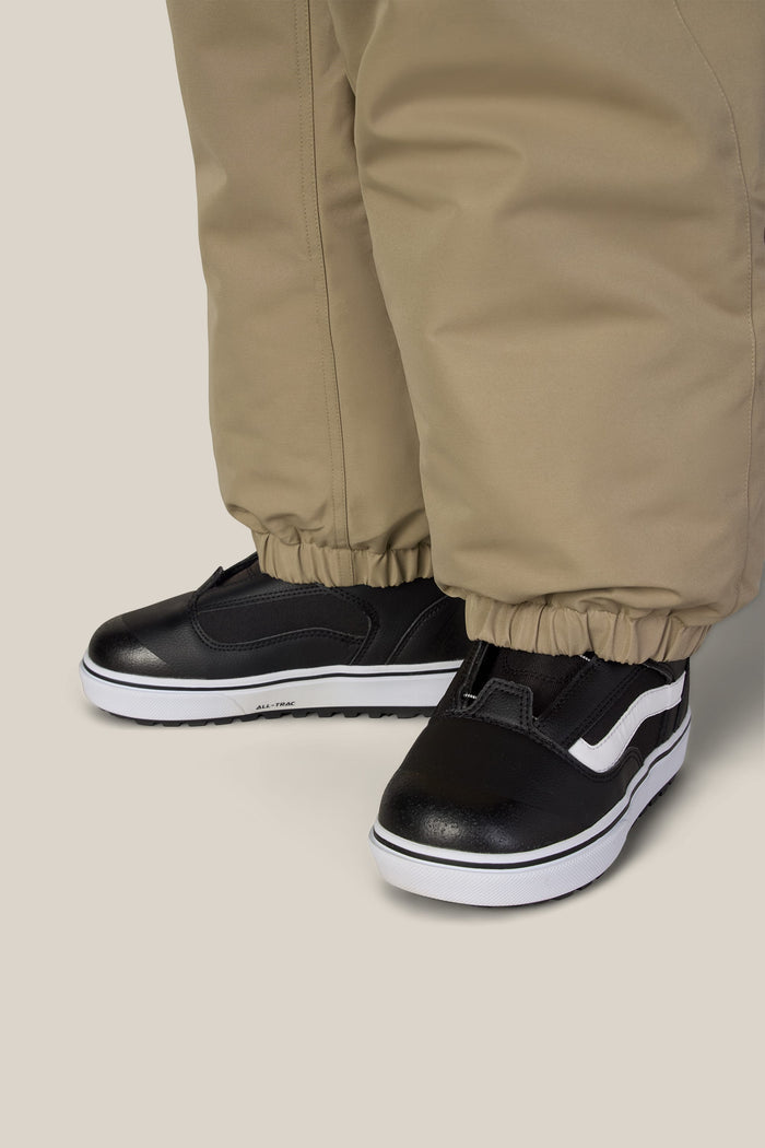 A child wearing 686 Boys’ Dojo® Insulated Pant in tan, featuring elastic cuffs, paired with black slip-on shoes with white soles and a white stripe on the side.