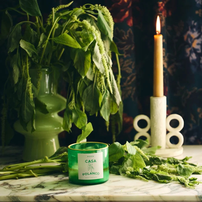Roen Casa Polanco candle in a green glass jar surrounded by fresh green verbena leaves and botanicals, styled with a lit taper candle on a marble surface