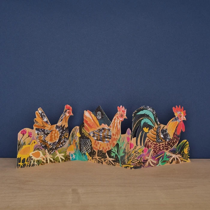 Decorative paper cutout card of chickens on a wooden surface with a blue background