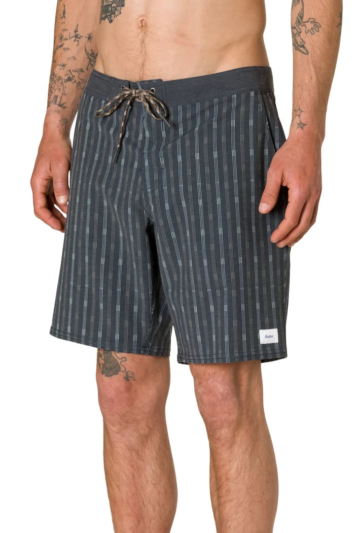Three-quarter front view of a tattooed man wearing the Katin Crossroads Hybrid Trunk in Black Wash, showing the vertical stripe print, lace-up waist with braided drawcord, and woven Katin label on the lower right leg