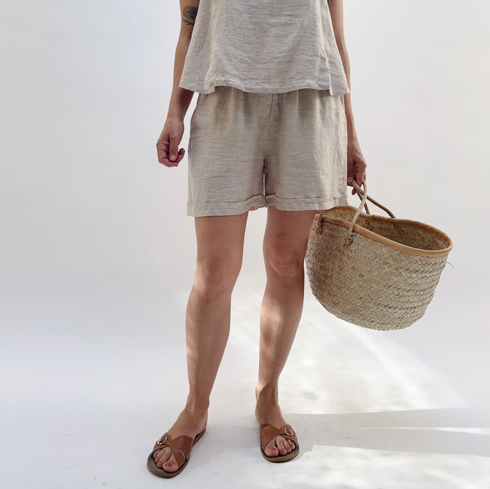 Person wearing a beige linen tank top and matching linen shorts holding a woven basket on a white background.