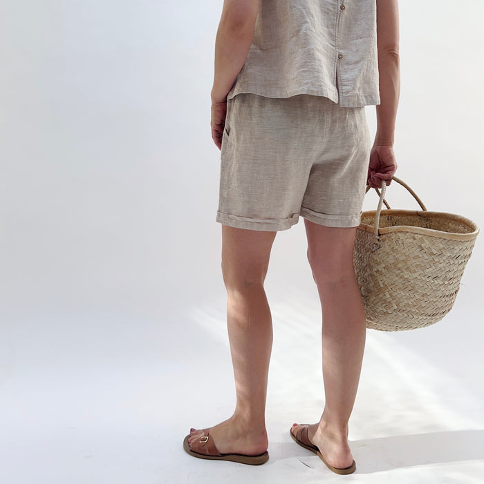 Person wearing a beige linen tank top and matching linen shorts holding a woven basket on a white background turned away from the camera.