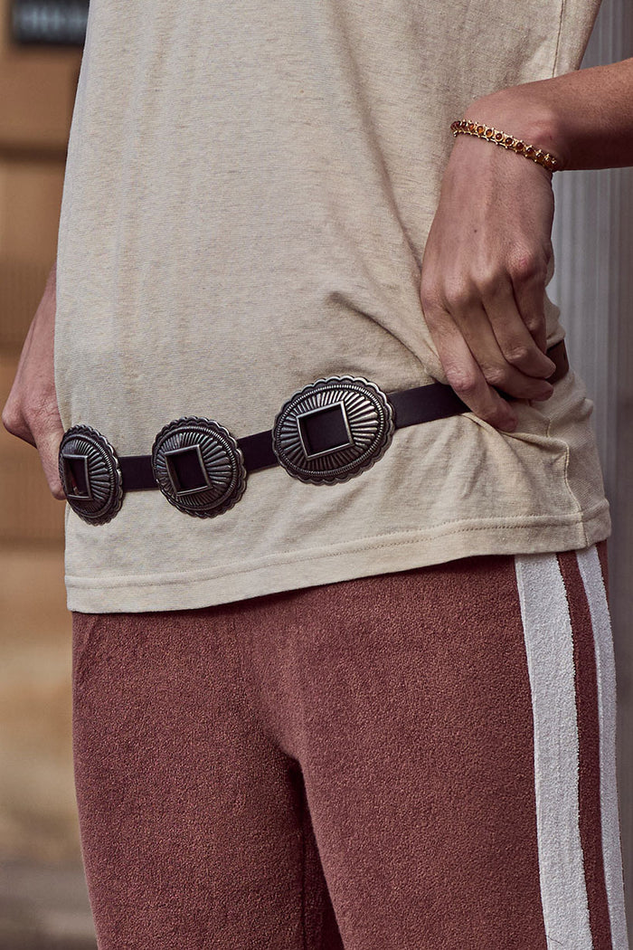 Close-up of a model wearing the Elysian Belt, dark leather with antique silver concho hardware styled over a tee