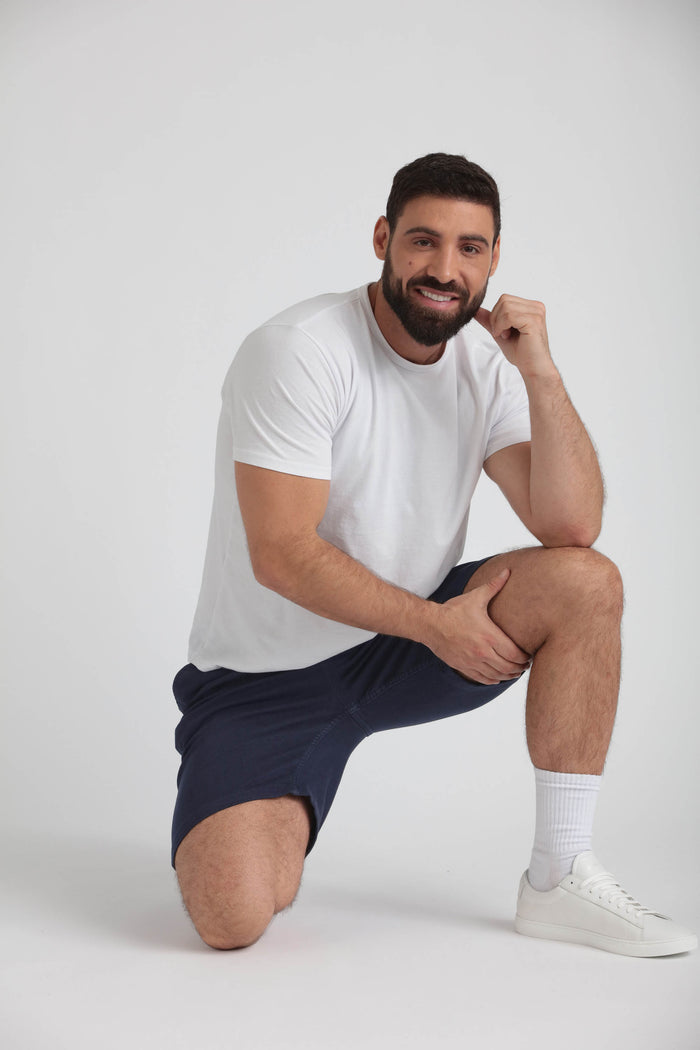 Man wearing white t-shirt and navy athletic shorts, kneeling against white background.