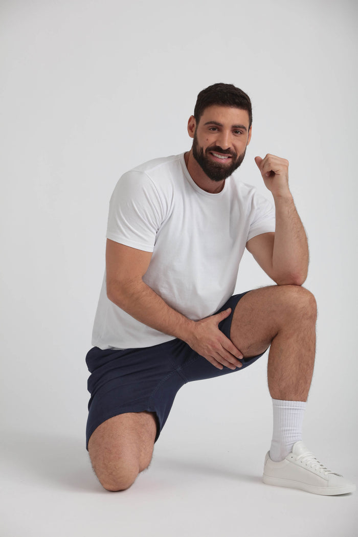 Man wearing white t-shirt and navy athletic shorts, kneeling against white background.