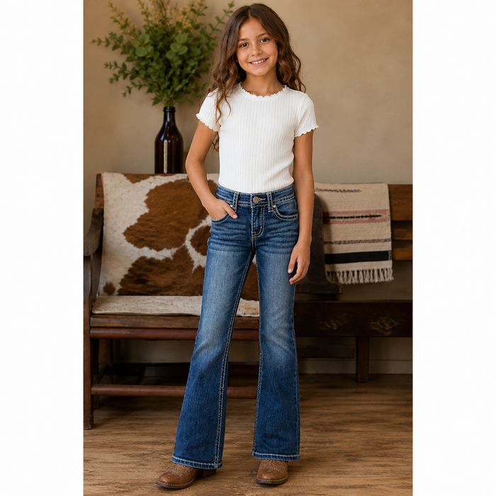 Young girl wearing a white top and blue jeans standing in a room with a wooden bench and decorative items.