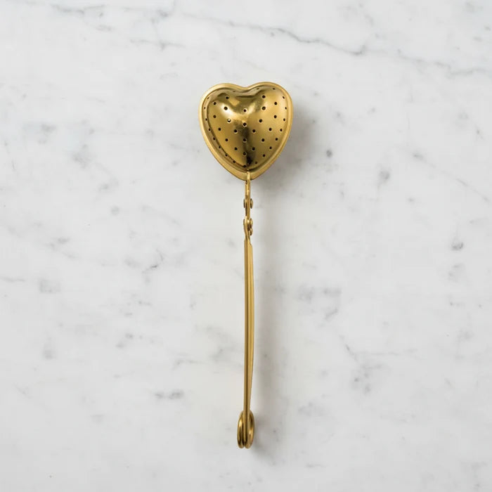 Gold-finish stainless steel heart-shaped tea strainer with squeeze handle, laid flat on a white marble surface