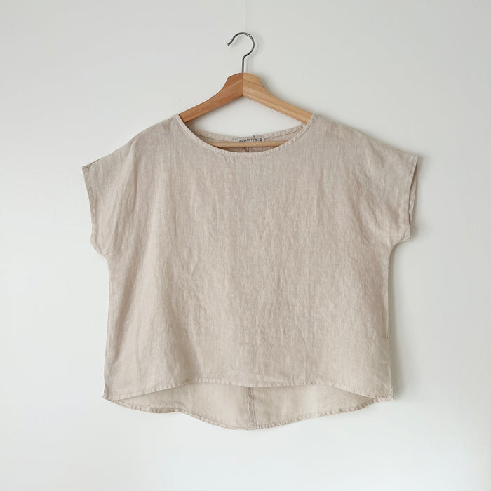 A beige high-low hem linen tee hanging on a wooden hanger against a white wall.