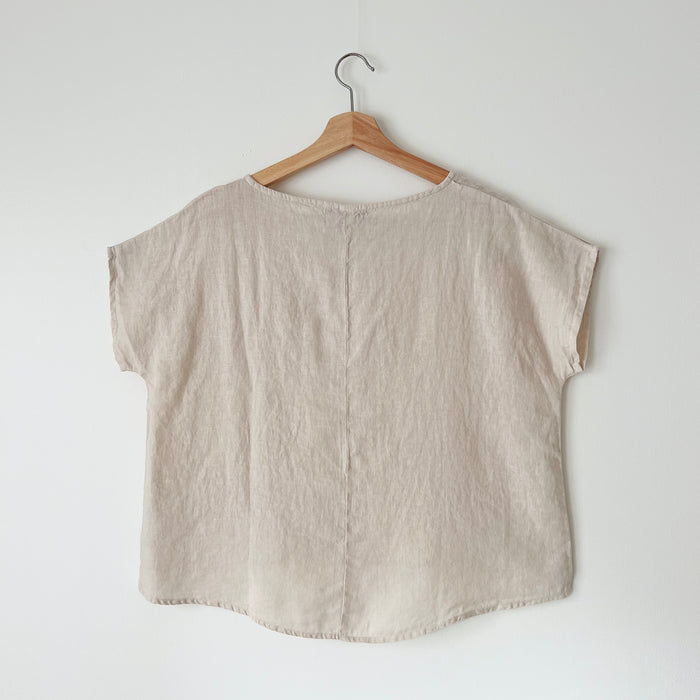 A beige high-low hem linen tee hanging on a wooden hanger against a white wall, viewed from the back