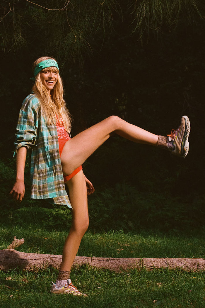 Side view of oversized flannel check shirt with dropped shoulder, styled open over orange swimwear, leg raised outdoors
