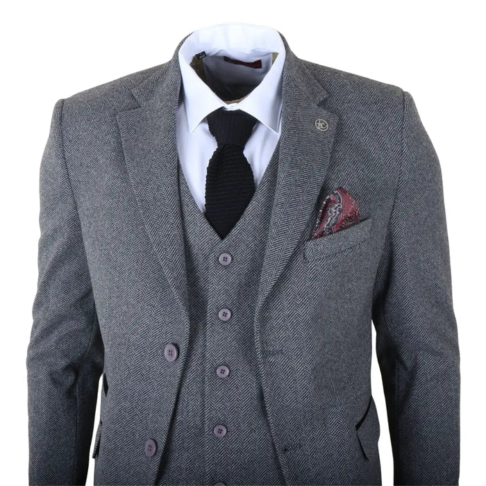 Men's grey tweed three-piece suit with a white shirt, black tie, and patterned pocket square, in a well-lit industrial workspace
