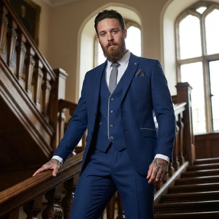 Man wearing a stylish blue tailored suit with a fitted blazer, vest, white shirt, grey tie, and tattooed hands, standing on a staircase in an elegant setting