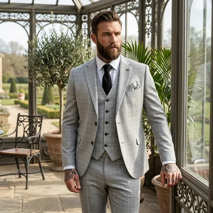 Man wearing light grey check suit with matching blazer and trousers, white shirt, black tie, and pocket square, standing in bright conservatory, SKU: IM53_suit_Grey_34