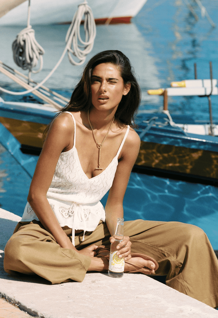 Model seated dockside in white pointelle cami, waist tie cinched, sweetheart neckline, paired with olive wide-leg pants