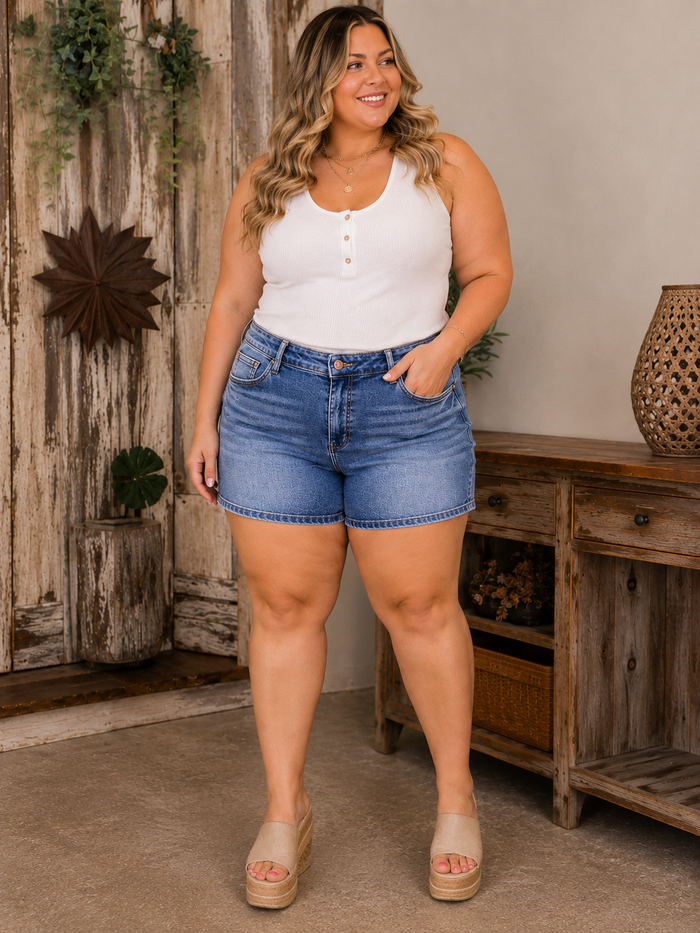 Woman wearing a white tank top and blue denim shorts by Judy Blue size 2XL standing in a room with wooden furniture.