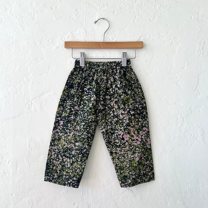 Kids lazy pants in green and pink speckle Batiki cotton, hanging flat on a wooden clip hanger against a white textured wall, showing elasticated waistband, dark navy ground with hand-printed green, pink, and white speckle pattern, and relaxed fit