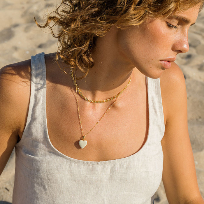 Person at the beach wearing a beige top and heart-shaped white locket and a shorter gold necklace