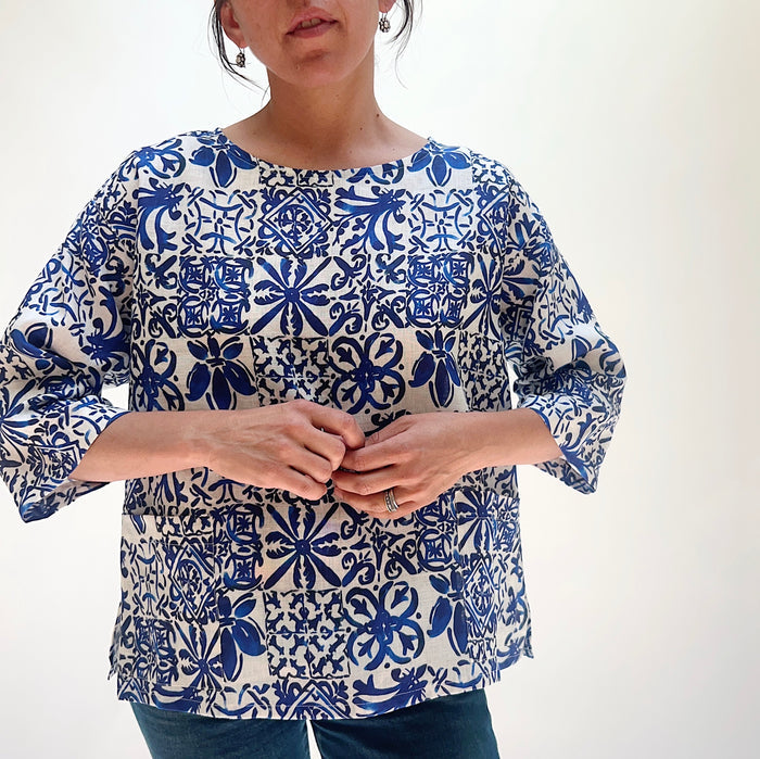 Close-up front view of the Christopher Calvin Linen Blue Tiles Print Top showing the blue-and-white Moroccan tile print, boat neckline, and 3/4 sleeves, worn with blue jeans on a white background