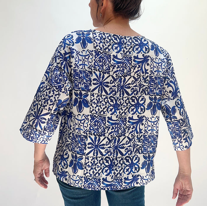 Rear view of the Christopher Calvin Linen Blue Tiles Print Top showing the blue-and-white tile print across the back, boat neckline, and 3/4 sleeves, worn with blue jeans on a white background